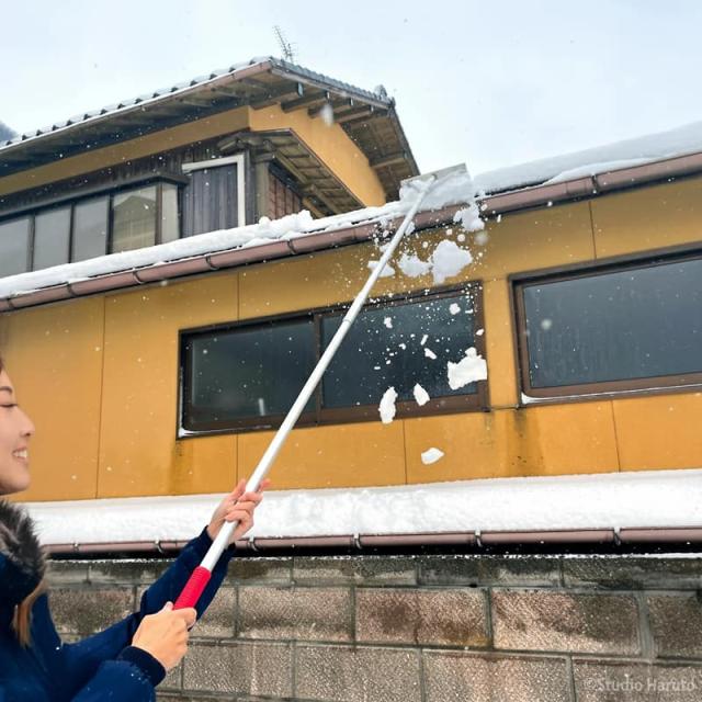 建物の屋根から、雪下ろし道具を使って雪を除去している様子。冬の住宅メンテナンスの一環として、積雪による被害を防ぐための作業風景を示している。