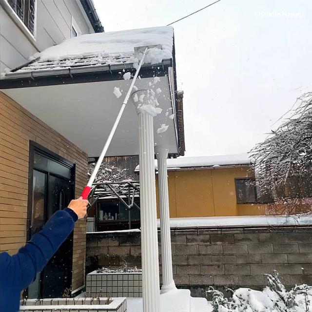 雪が積もった住宅の屋根から、長柄の雪下ろし道具を使って雪を除去している様子。屋根の端から雪を掻き出している。
